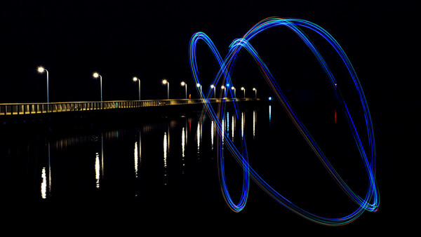 Long exposure photograph of a pier at night with blue light trails spiraling from its end.