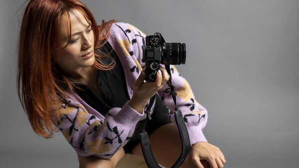 Woman holding a film camera to her eye, wearing a lavender patterned cardigan against a neutral gray background.