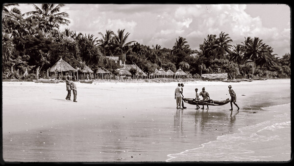 Multiple figures launching a wooden boat into shallow ocean waters with palm-lined beach village visible in background.
