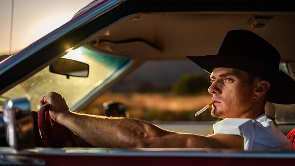 Man in cowboy hat smoking cigarette while driving vintage car at sunset.