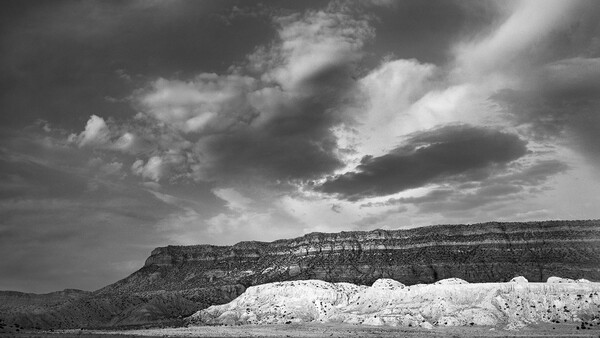 Dramatic black and white landscape with layered mesa formations and white cliff face under stormy sky.