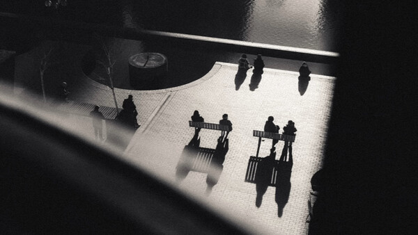 Overhead view of silhouetted figures casting long shadows on a bright floor beneath architectural elements.