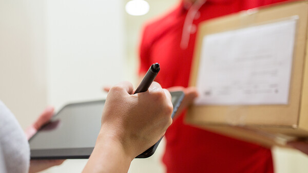 Hand holding a stylus pen with a blurred whiteboard and red fabric in the background.