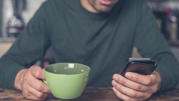 Person holding a green mug and smartphone while seated at a table, wearing a dark long-sleeved shirt.