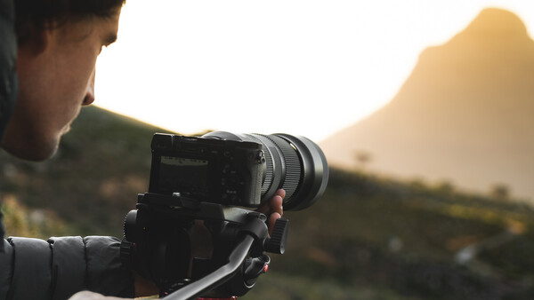 Photographer operating a mirrorless camera on a tripod during golden hour with mountains in the background.