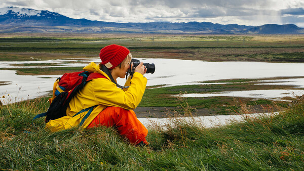 Photographer in yellow jacket and red hat crouching while photographing a river valley landscape with mountains.