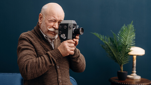 Older man with beard holding vintage film camera up to his face against blue background.