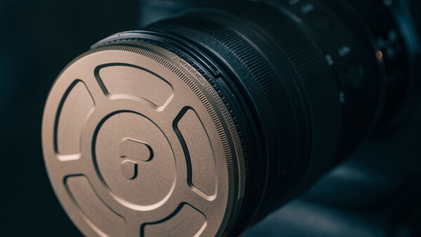 Close-up of a metallic lens cap with circular venting slots and center post, mounted on a camera lens barrel.