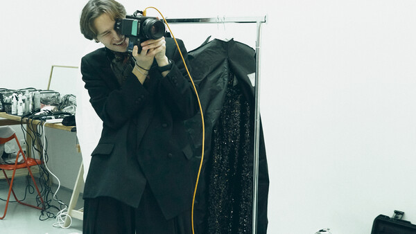 Photographer holding a camera to their eye while standing next to a clothing rack with a dark garment.