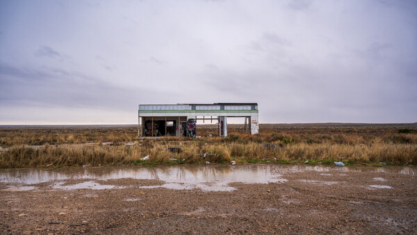 Abandoned single-story building with metal siding in vast, flat marshland under overcast sky.