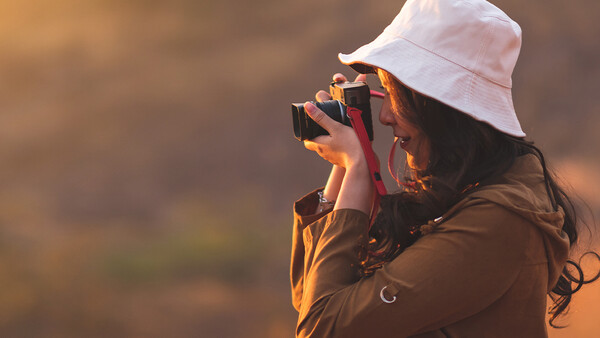 Photographer with white bucket hat holding camera to face, backlit by warm golden light against blurred background.