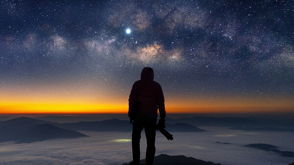 Silhouetted photographer standing on a mountain peak overlooking clouds at sunrise, with the Milky Way and stars visible in the night sky above.