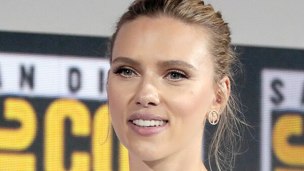 Woman with blonde hair pulled back, smiling at camera against blurred yellow and black signage background.