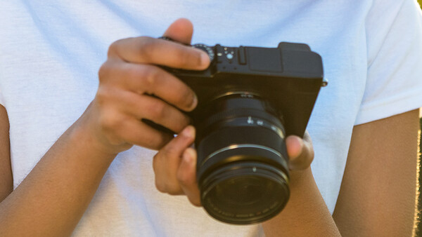 Photographer holding a mirrorless camera with attached lens against a light background.