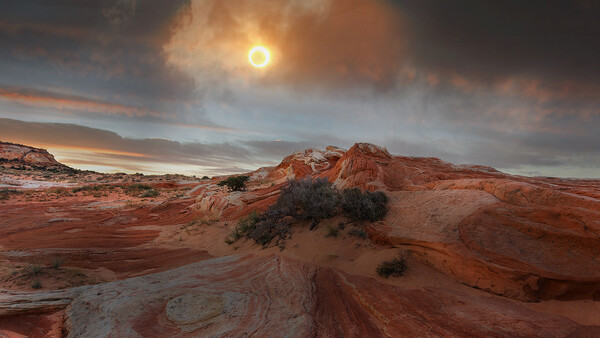 Desert landscape with layered red rock formations beneath a dramatic golden-hour sky with sun breaking through clouds.