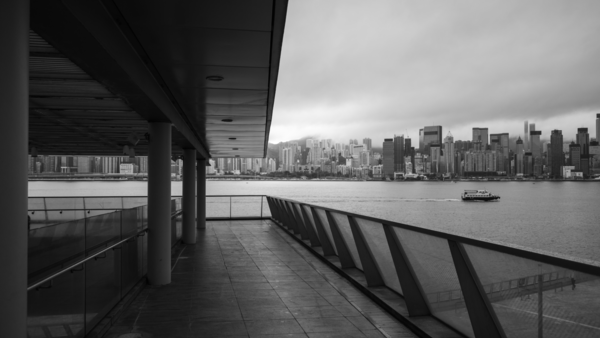 Black and white view from a covered waterfront pier toward a distant city skyline across calm water.