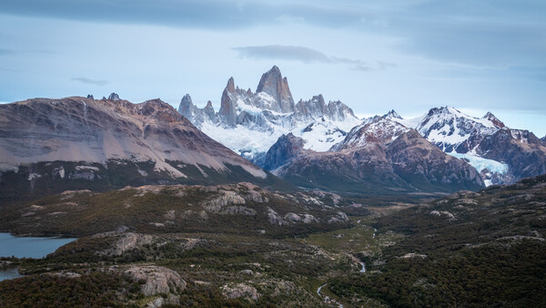 Snow-capped mountain peaks with jagged granite formations rising above a turquoise glacial lake and forested foothills.