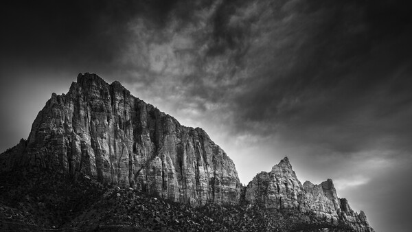 Dramatic black and white photograph of steep columnar rock formations rising against a turbulent sky.