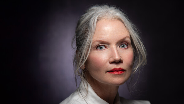 Portrait of a woman with gray hair and red lipstick against a dark background.