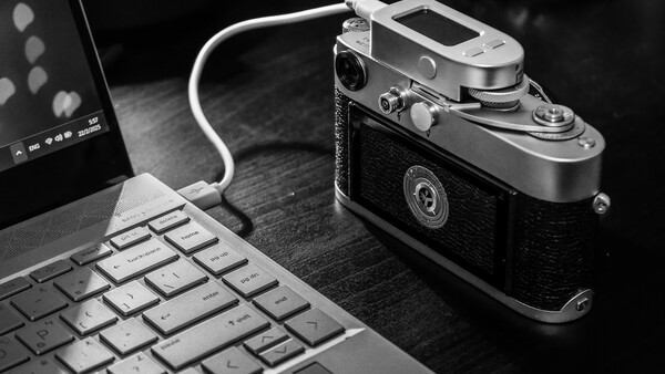 Rangefinder film camera connected to laptop via USB cable on wooden desk.