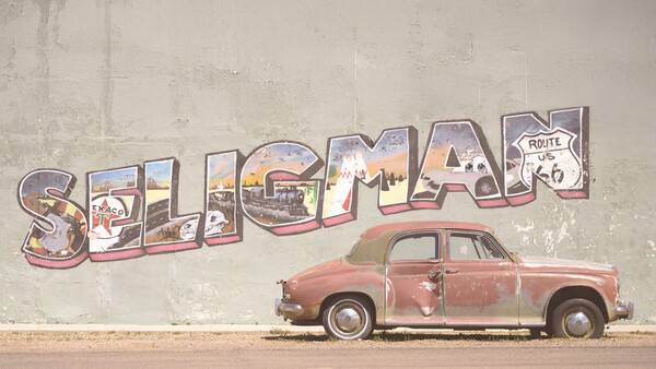 Vintage red car parked in front of weathered wall mural spelling 'Seligman' with Route 66 imagery.