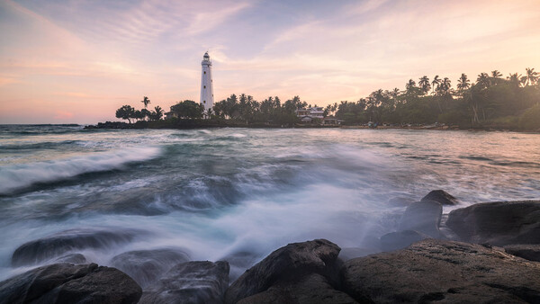 Lighthouse on a peninsula with palm trees, photographed from a rocky beach at golden hour with waves in the foreground.