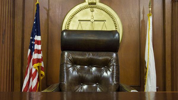 Empty judge's chair in a courtroom with scales of justice symbol behind it.