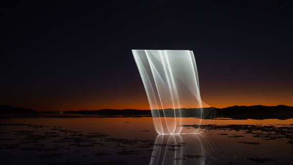 Light painting over a still body of water at dusk with warm orange sky and dark shoreline silhouettes.