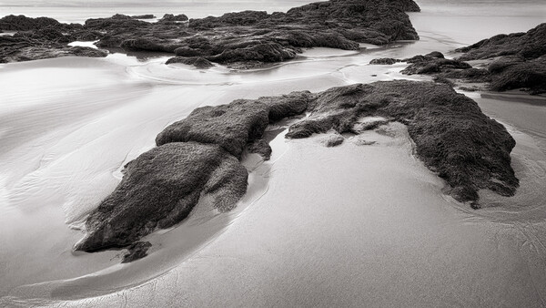 Rocky outcrops emerging from smooth sand on a misty beach with long-exposure water blur.