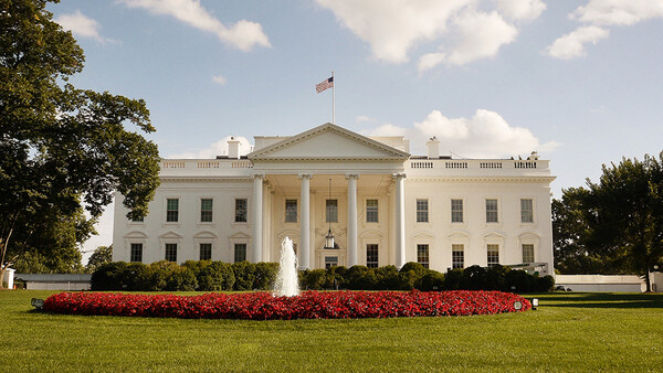 White neoclassical building with columned facade, manicured lawn, and red flowering plants in foreground.