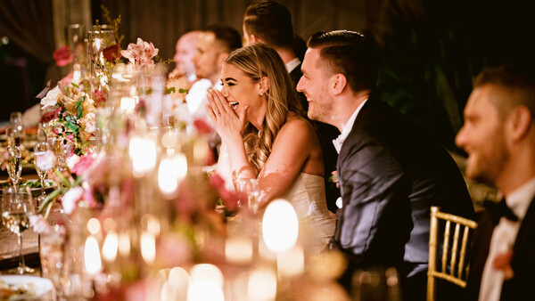 Bride and groom laughing together at an elegantly decorated wedding reception table with pink floral arrangements and candlelight.