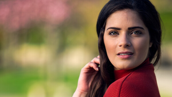 Woman with dark hair wearing a red sweater, looking at camera with soft expression against blurred background.