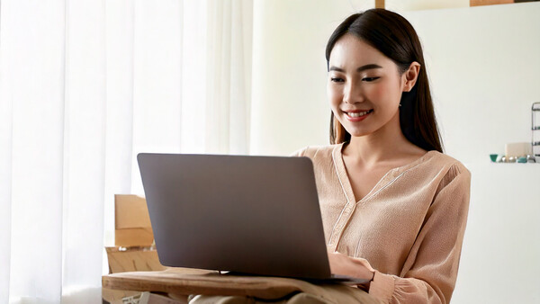 Woman in beige cardigan smiling while working on laptop at desk with cardboard boxes.
