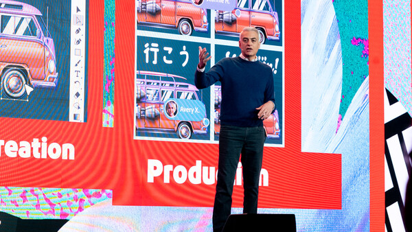 Speaker on stage in front of large red screen displaying colorful vintage cars and production text.
