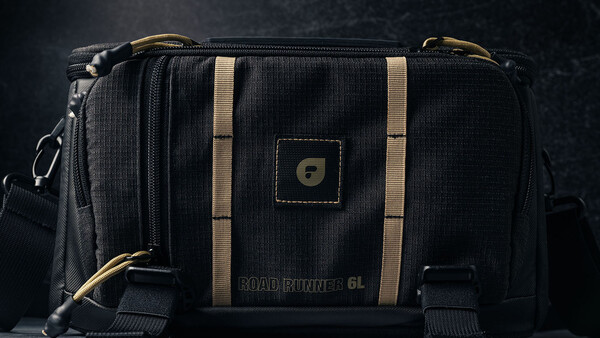 Black camera bag with tan stripes and brass hardware against dark background.