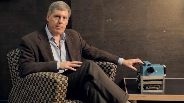 Man in business casual attire seated in patterned chair next to vintage projector.