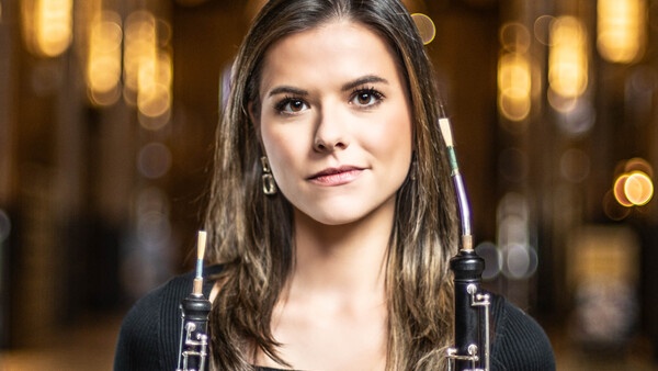 Portrait of a young woman holding oboes against a warm, blurred interior background.