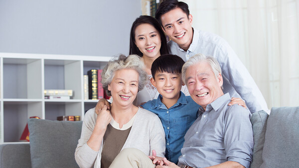 Multi-generational Asian family of five smiling together on a gray couch in a modern living room.