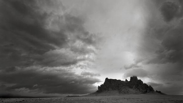 Silhouetted rocky pinnacles rise from a flat desert plain beneath a dramatic storm sky.