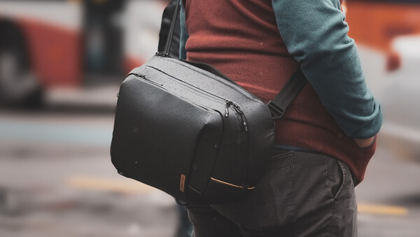 Person wearing a black camera backpack with shoulder strap over casual clothing in an urban street setting.