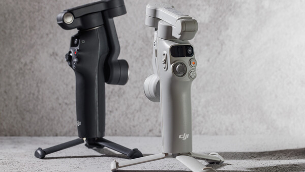 Two smartphone gimbals displayed side by side on a concrete surface, one black on the left and one white/gray on the right.