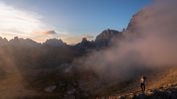 Hiker standing on a rocky slope with dramatic mountain peaks and misty valleys at golden hour.
