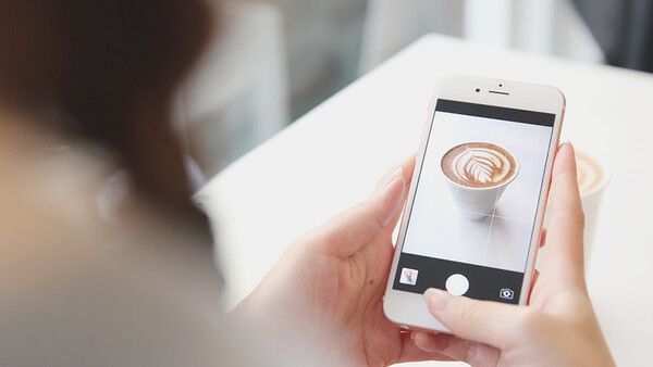 Person holding white smartphone displaying close-up photo of latte art in camera app.