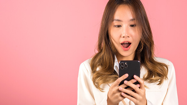 Woman in white shirt holding smartphone with surprised expression against pink background.