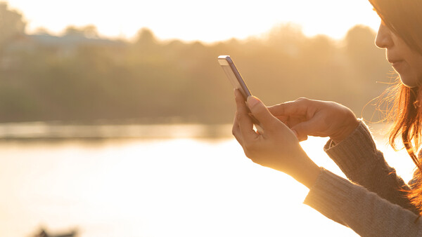 Woman holding smartphone toward water during golden hour, capturing landscape with backlighting.