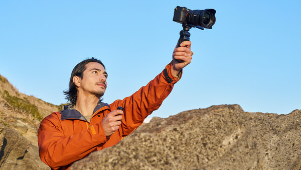 Man in orange jacket holding smartphone on gimbal stabilizer against desert hillside.