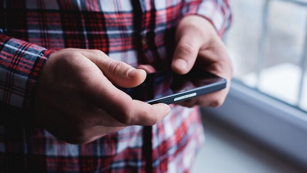 Close-up of hands holding a smartphone with a plaid shirt visible, shot near a window with natural lighting.