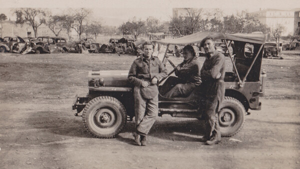 Three men in military uniforms standing beside a utility vehicle in an open field with vehicles and structures visible in the background.