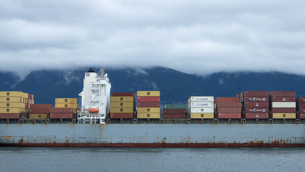 Large cargo ship stacked with colorful shipping containers against a mountainous coastline.