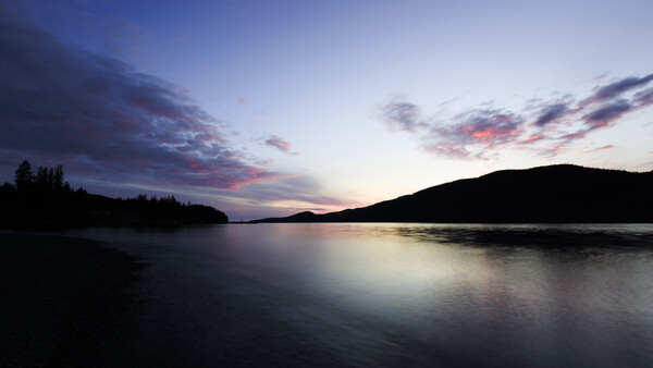 Twilight landscape with calm lake, forested shoreline, and mountains silhouetted against purple and pink sky.